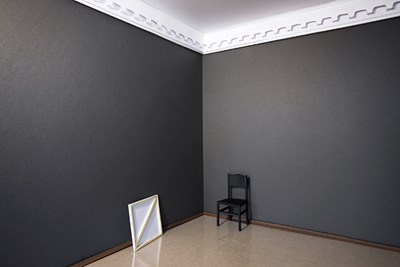 An empty room with a blank canvas leaning against one wall and a single black chair against the other