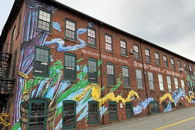 A mural celebrating the arts on the side of a former mill building that now houses the Essex Art Center, the Acting Out! Theatre Co. and Spicket River Brewery.